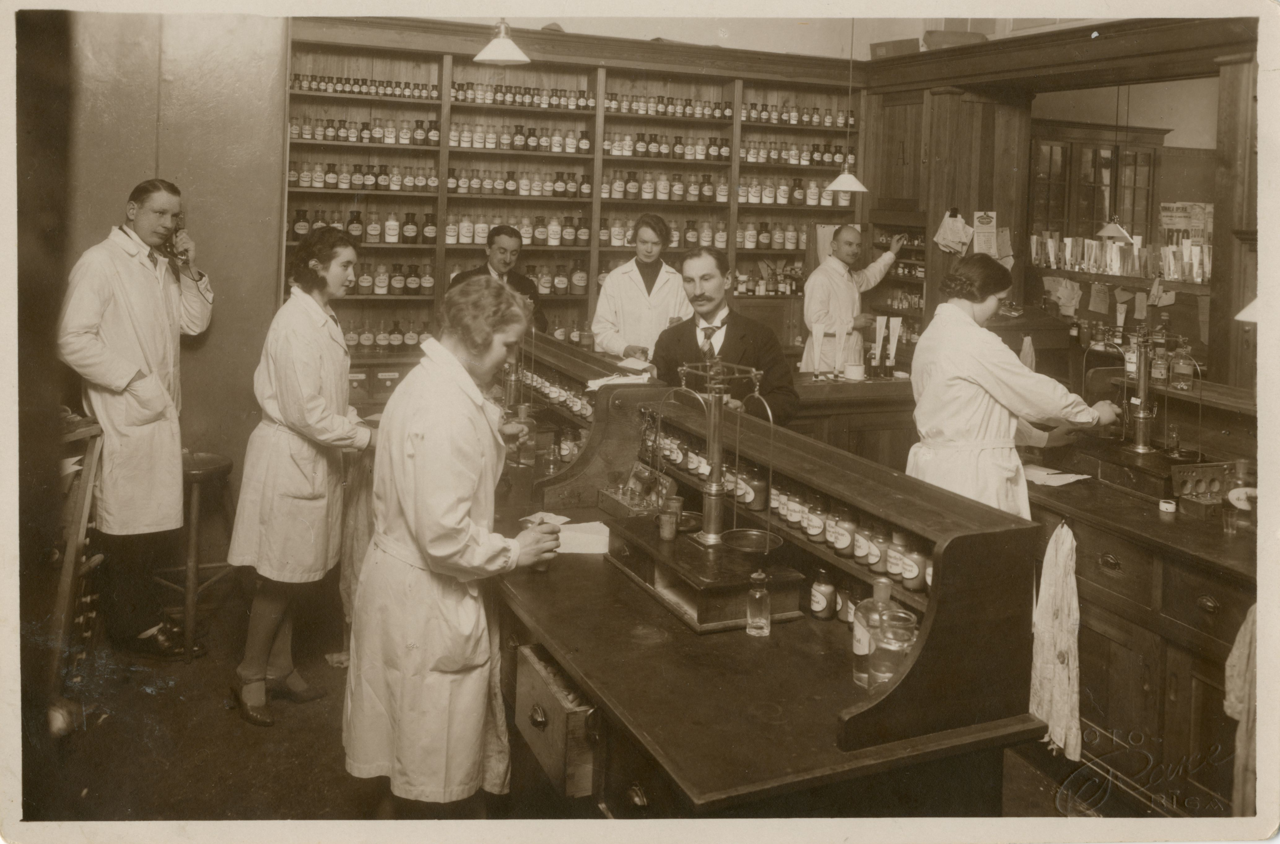 LU aptiekas asistentu telpa. Centrā pārvaldnieks H. Freijs, kreisajā pusē farmācijas maģistrs Artūrs Neilands. Paula Stradiņa Medicīnas vēstures muzeja krājums, MVM 29207 Ff 3746